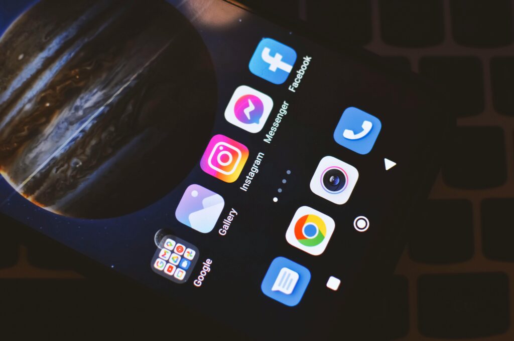 A close-up, angled view of a smartphone screen displaying social media and utility apps, set against a planet-themed wallpaper and placed over a laptop keyboard.