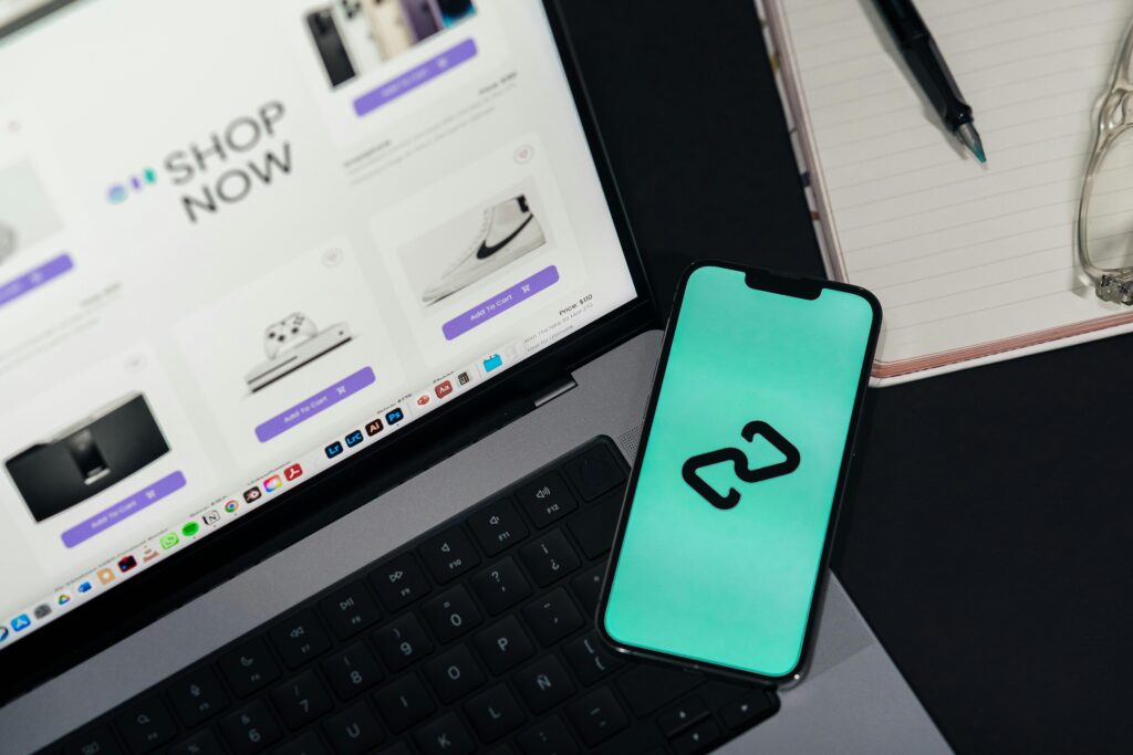 A smartphone with a green screen displaying a black abstract logo lies on a laptop keyboard next to an open e-commerce website showing "SHOP NOW."