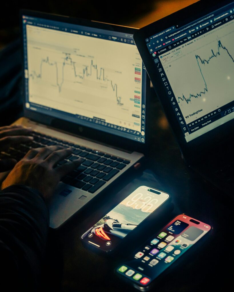 A person typing on a laptop in a dimly lit room, with financial stock charts displayed on two laptop screens. Two illuminated smartphones sit on the desk in the foreground.